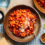 A big bowl of the pasta with chickpeas, sitting on a tabletop alongside a block of parmesan, drink glasses, and another bowl of the pasts