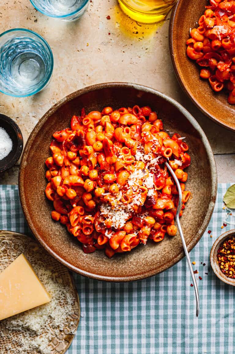 A big bowl of the pasta with chickpeas, sitting on a tabletop alongside a block of parmesan, drink glasses, and another bowl of the pasts