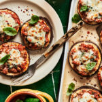 A plate of the mini eggplant pizzas, sitting on a countertop next to a tray of more of the pizzas
