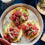 A plate of 3 of the ahi tuna tostadas, sitting on a plate one a countertop surrounded by a margarita glass and a bowl of tortillas