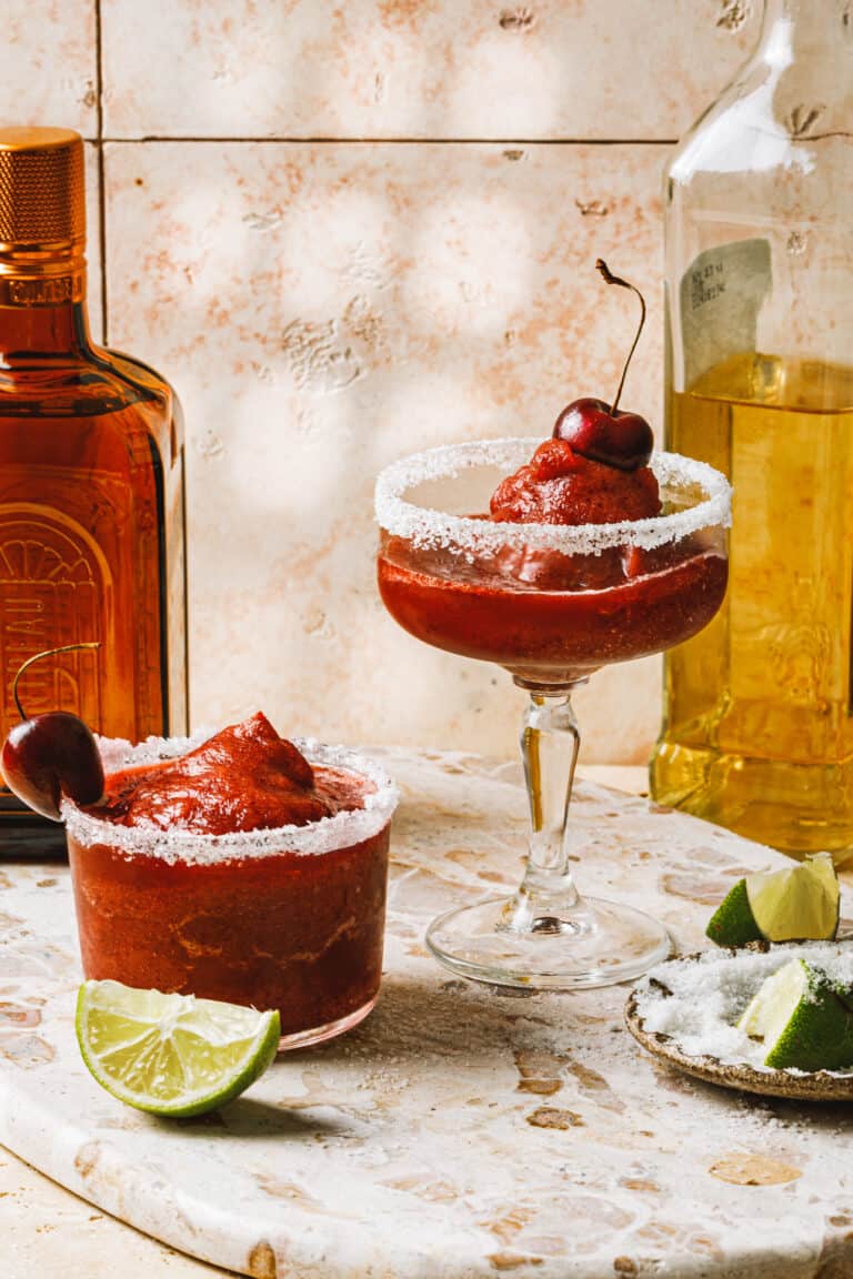 Two tall glasses of the frozen cherry margaritas, toped with cherries and sitting on a countertop next to a wedge of lime