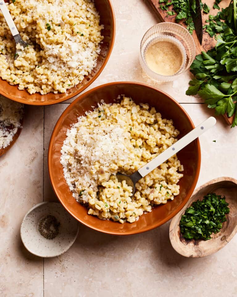 A bowl of the grown-up alphabet pasta, sitting in a bowl on a countertop. A larger bowl of the pasta and a small plate of parsley sits next to the bowl