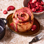 One of the single serving strawberry cinnamon rolls, on a plate, with fresh strawberries in a bowl behind it. A cast iron ramekin can be seen off to the side.