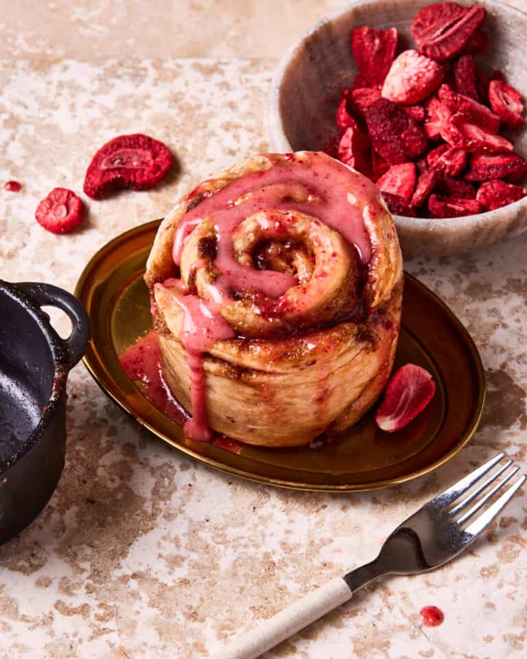 One of the single serving strawberry cinnamon rolls, on a plate, with fresh strawberries in a bowl behind it. A cast iron ramekin can be seen off to the side.