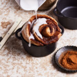 A cast iron ramekin, filled with one of the single serving cinnamon rolls, with the icing being actively poured on top of it
