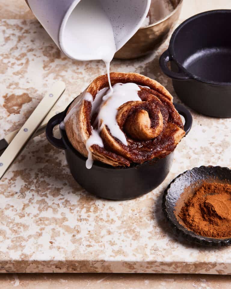A cast iron ramekin, filled with one of the single serving cinnamon rolls, with the icing being actively poured on top of it