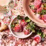 A bowl of the drunken strawberries, sitting on a countertop next to a glass of wine and a larger bowl of strawberries