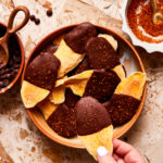 A plate of the chocolate dipped mango with tajin, sitting on a countertop with a hand reaching for one of the slices