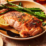 A close-up image of the perfect 8-minute broiled salmon, sitting on a plate alongside asparagus spears and a lemon wedge