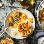 A plate, topped with bulbs of roasted and smashed garlic, sitting on a countertop next to more heads of garlic and a bottle of olive oil