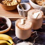 Two of the gut healthy creamy date cashew shakes, sitting on a countertop, with bananas and cashews in the background