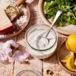 A bowl of the healthy Caesar salad dressing, sitting on a countertop, surrounded by different ingredients needed to make the dressing