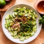 A bowl of the spicy cucumber & avocado salad, sitting on a countertop with other ingredients stacked in the background