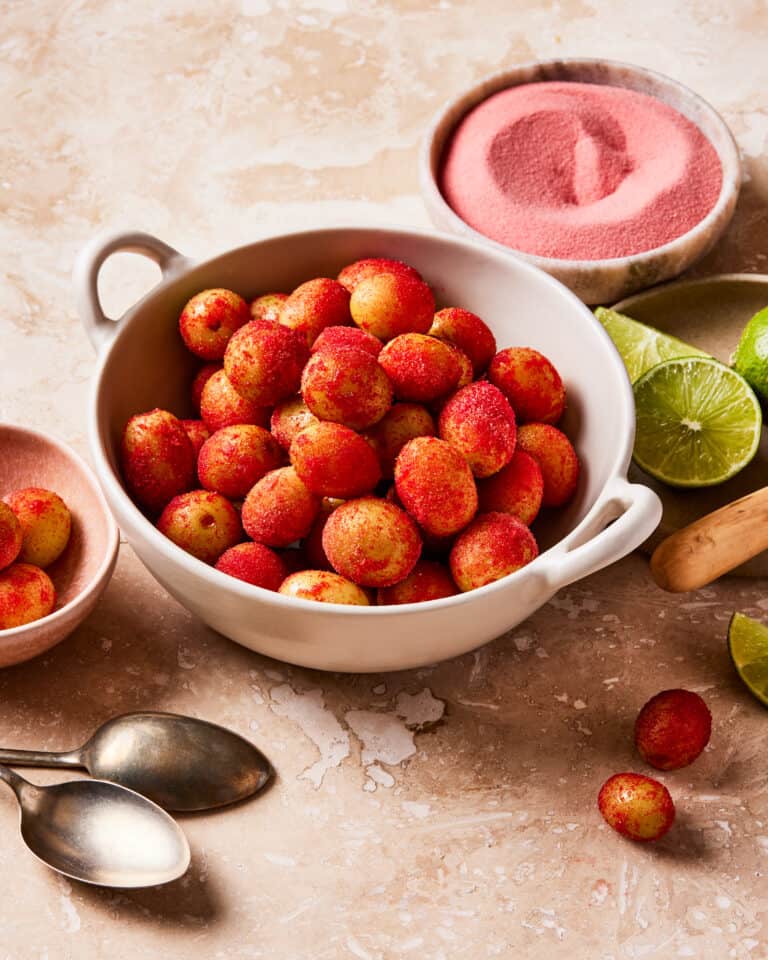 A bowl of the sour patch strawberry frozen grapes, sitting on a countertop next to spoons, cut lime wedges, and a bowl of jell-o mix