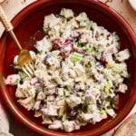 A red bowl, sitting on a countertop, with the mixed up Ultimate Healthy Chicken Salad sitting inside of it. A gold fork can also be seen sitting inside of the bowl, sticking out of a bite of the chicken salad