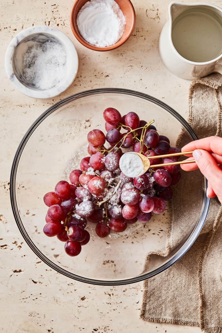 A clear bowl with the grapes inside of it, and a hand pouring the sea salt and baking soda on top of the grapes.