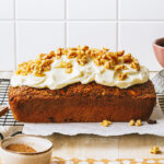 The one bowl healthy carrot cake loaf, sitting on top of a wire cooling rack, and topped with cream cheese frosting and chopped walnuts