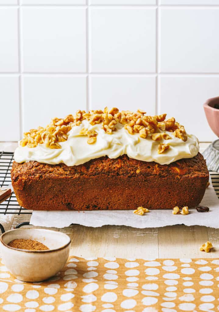The one bowl healthy carrot cake loaf, sitting on top of a wire cooling rack, and topped with cream cheese frosting and chopped walnuts