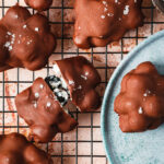 A close-up image of the blueberry yogurt clusters, sitting on a wire cooling rack alongside a teal plate with one of the clusters on top of it