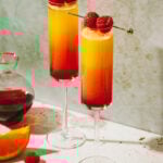 Two glasses of the ombre tequila mimosa, sitting on a countertop next to orange wedges and a bottle of grenadine