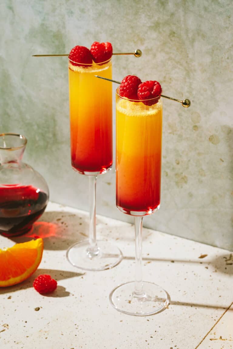 Two glasses of the ombre tequila mimosa, sitting on a countertop next to orange wedges and a bottle of grenadine