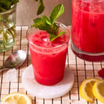 A glass of the five minute raspberry lemonade refresher, topped with a sprig of mint, and sitting on a countertop next to slices of lemons.