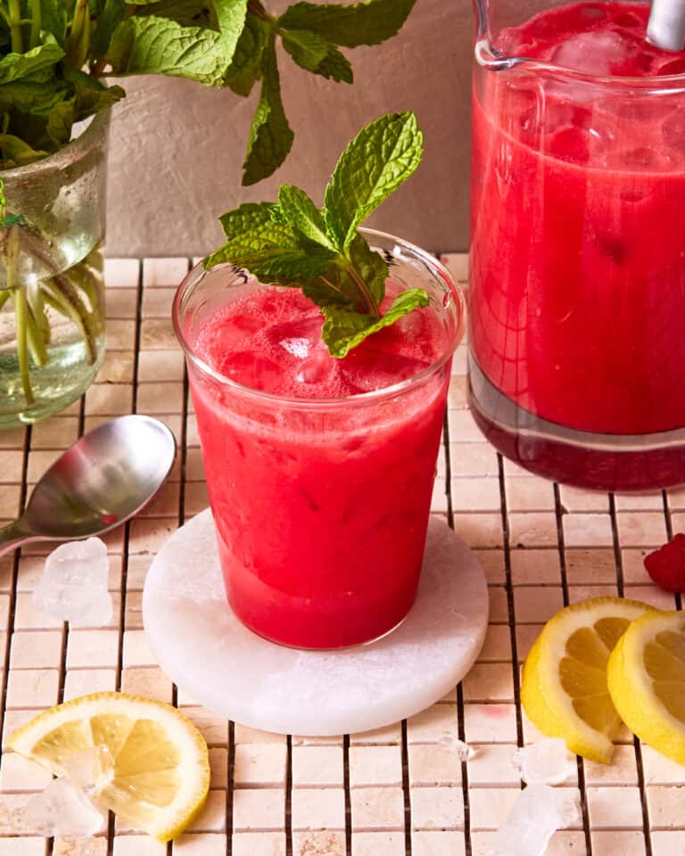 A glass of the five minute raspberry lemonade refresher, topped with a sprig of mint, and sitting on a countertop next to slices of lemons.