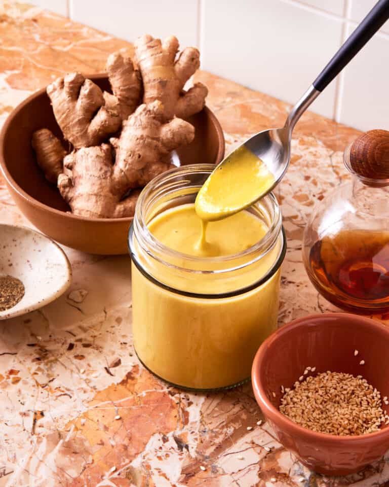 A jar of the Miso carrot dressing, sitting on a countertop alongside a bowl of ginger. A spoon is scooping the dressing out of the jar