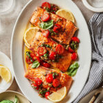 A plate of salmon with the tomato lemon sauce, sitting on a countertop surrounded by napkins, utensils, and glasses of water