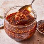 A jar of the Chili Onion Crunch, sitting on a countertop, with a spoon scooping some of the seasoning out of it
