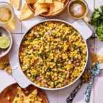 A bowl of the copycat chipotle corn salad, sitting on a countertop alongside a cutting board and a bowl of tortilla chips