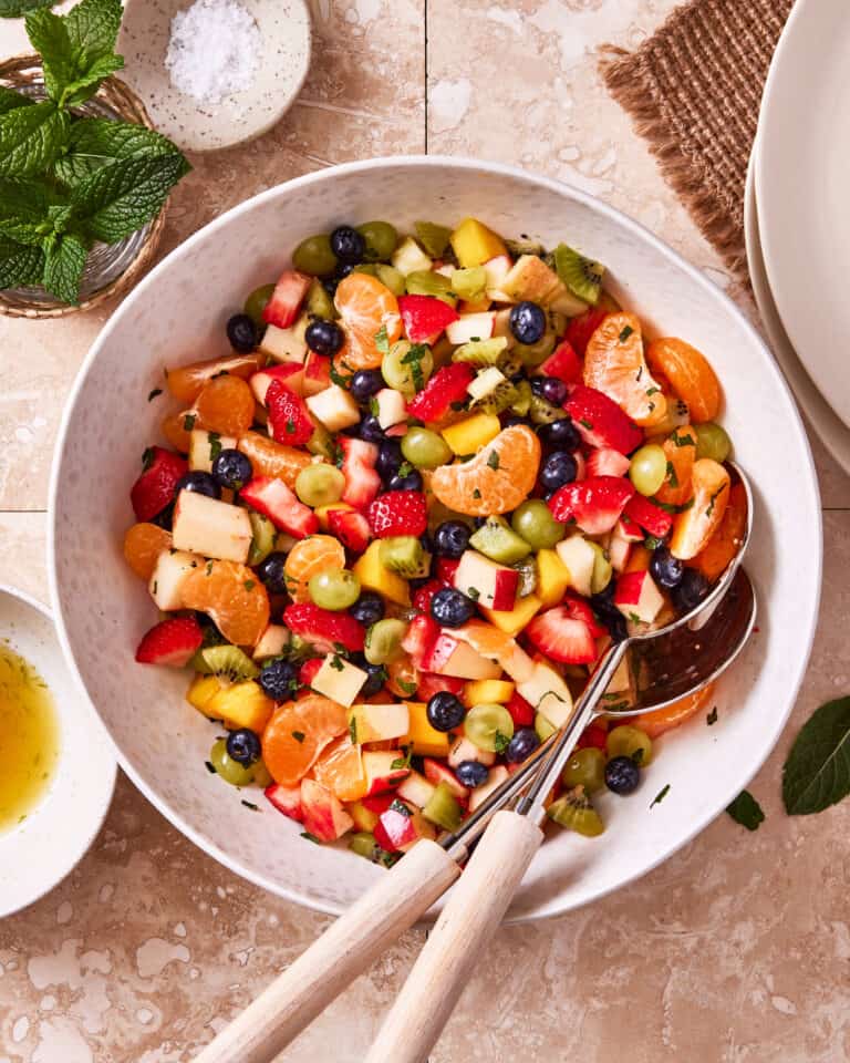 A large bowl of the ultimate fruit salad, sitting on a countertop, with serving spoons sitting inside of the bowl