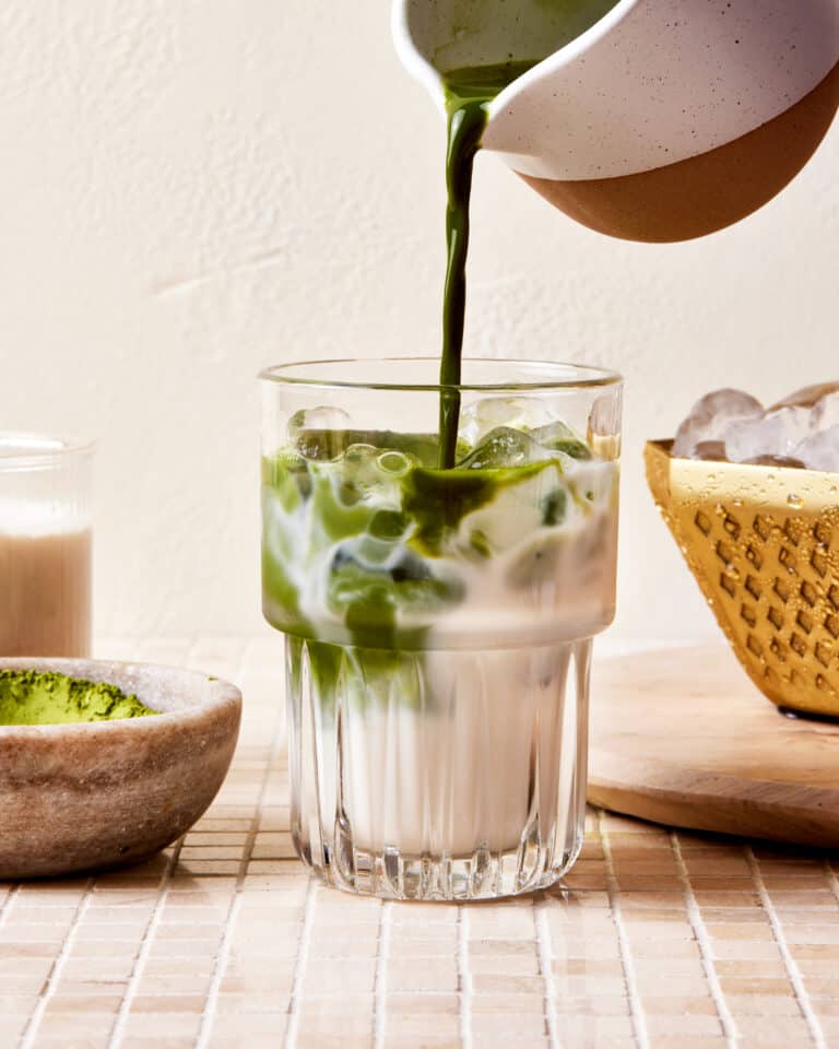 A glass, filled with homemade oat milk and ice, with the matcha latte actively being poured into it