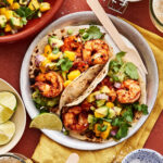 A plate of the air fryer shrimp tacos, sitting on a countertop surrounded by a bowl of salsa and a small bowl of guacamole