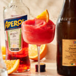 A tall glass of the blended aperol spritz, sitting on a countertop next to bottles of aperol and Prosecco