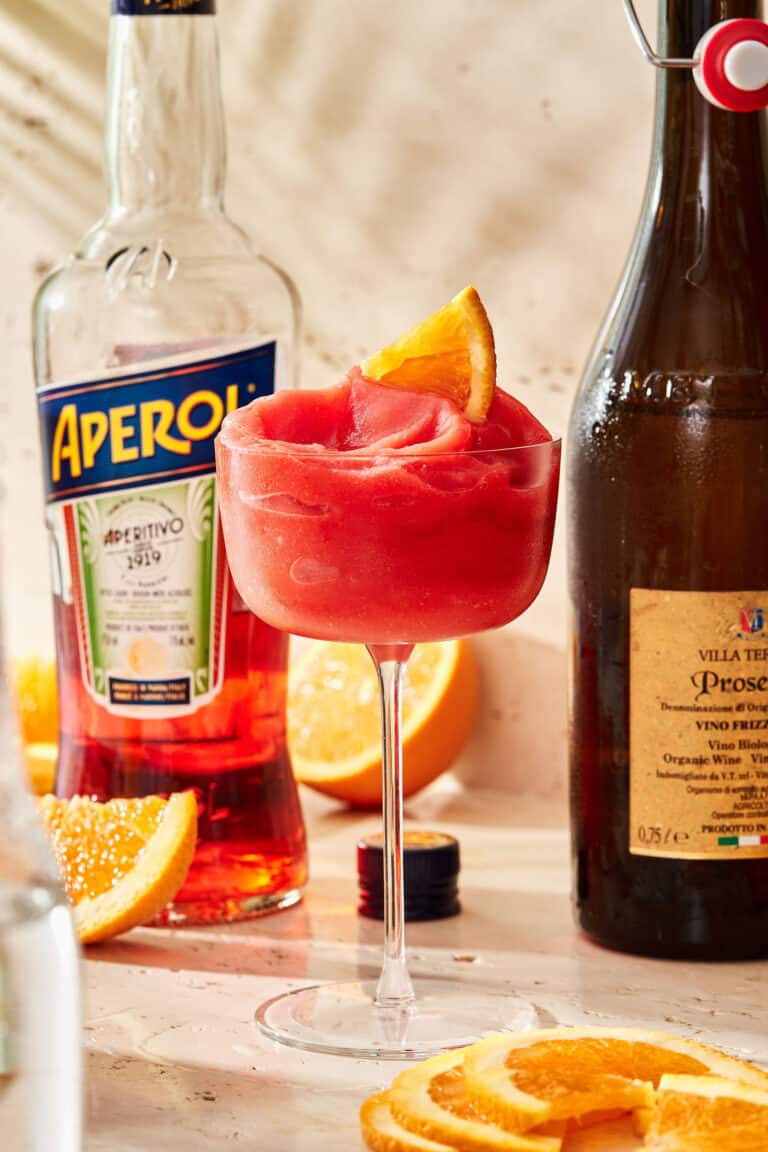 A tall glass of the blended aperol spritz, sitting on a countertop next to bottles of aperol and Prosecco