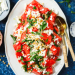 A plate of the watermelon feta salad, sitting on a countertop with serving utensils inside of it