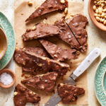 The snickers crunch bark, sitting on a piece of parchment paper next to a small bowl of raw peanuts and a knife