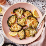 A bowl of the lemon parmesan smashed broccoli, sitting on a countertop next to a bowl of lemon wedges and a small bowl of parmesan cheese