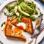 A plate, topped with the grilled cheese egg in a hole, next to an arugula and avocado salad