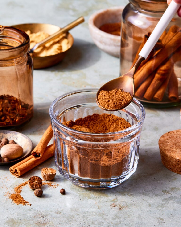 A jar of the homemade pumpkin pie spice, sitting on a countertop with a spoon scooping some of the spice out of the jar