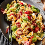 A bowl of the BLTE pasta salad, sitting in a bowl on a countertop with a serving fork and spoon sitting inside of it