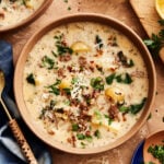 A bowl of the hearty Zuppa Toscana, sitting on a countertop surrounded by ingredients and a larger pot of soup