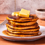 A stack of protein pancakes, sitting on a countertop with butter and maple syrup actively being poured on top of them