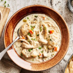 A bowl of the chicken dumpling soup, sitting on a countertop with a spoon resting on the edge of the bowl