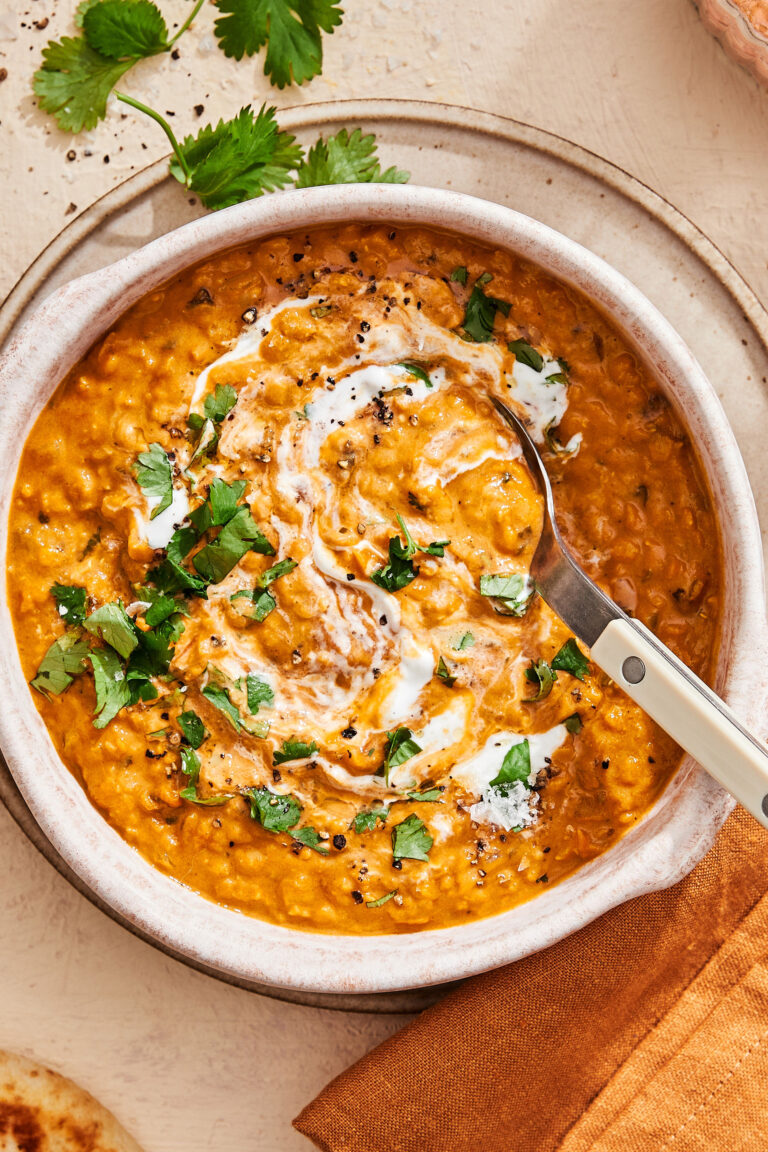 A close-up image of a bowl of the one pot curry lentil soup, sitting on a countertop topped with chopped cilantro and greek yogurt. A spoon is sitting inside of the bowl