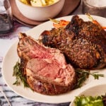 A close-up image of the ultimate easy prime rib, sitting on a plate on a countertop. A small bowl of mashed potatoes can be seen behind the plate.