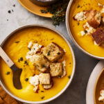 A bowl of the butternut squash feta soup, sitting on a countertop next to a larger pot of soup and a small ramekin of sea salt