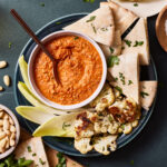 A plate of the romesco sauce, sitting on a countertop with a spoon sitting in the bowl and crudités around the sauce