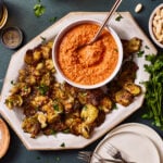 A plate of the crispy smashed Brussels sprouts with the creamy romesco sauce, sitting on a countertop with a spoon sitting in the sauce